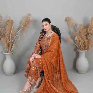 May include: A burnt orange three-piece ensemble featuring a long-sleeved tunic and matching trousers with intricate gold embroidery, sequins, and beadwork. A sheer, lightweight dupatta with a delicate gold geometric pattern drapes over the outfit.
