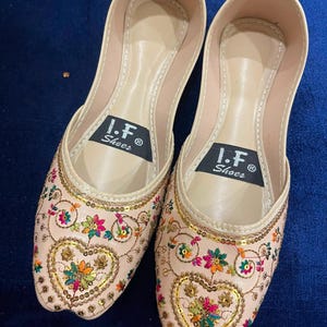 May include: A pair of light beige slip-on flats featuring intricate multi-colored floral embroidery and gold sequin detailing. The interior soles are lined in beige and feature a black label with the white text I.F. Shoes printed on each shoe.