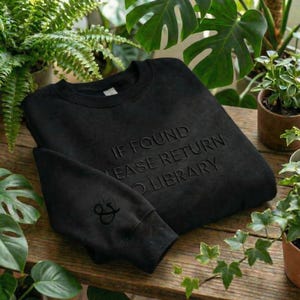 May include: Black sweatshirt with the embossed text "IF FOUND PLEASE RETURN TO LIBRARY." The sleeve features a floral and ampersand design. The sweatshirt is displayed on a wooden surface surrounded by potted plants.