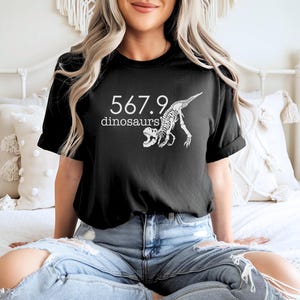 May include: Black t-shirt with white text and a dinosaur skeleton graphic. The text reads "567.9 dinosaurs". The shirt is a short-sleeved crew neck.
