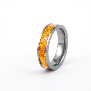 May include: A polished dark grey metal ring featuring a central inlay of crushed orange and yellow stone fragments. The band has a smooth, reflective surface and a flat profile, showcasing a vibrant, textured inlay that runs around the circumference.