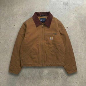 Puede incluir: Una chaqueta de lona color canela con cuello de pana marrón oscuro, cremallera frontal completa y dos bolsillos delanteros inclinados. Una pequeña etiqueta rectangular de la marca Carhartt está cosida en el bolsillo izquierdo del pecho y el cuello.