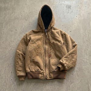 Peut inclure: Une veste à capuche beige avec une fermeture éclair frontale, une doublure intérieure matelassée noire, ainsi que des poignets et un ourlet côtelés. Le vêtement comprend deux poches avant et une petite étiquette rectangulaire de marque.