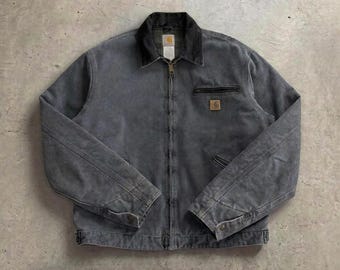 May include: A charcoal grey canvas work jacket featuring a dark grey corduroy collar, a full-length front zipper, and a zippered chest pocket. A small square Carhartt brand patch is stitched onto the left chest area above the pocket.