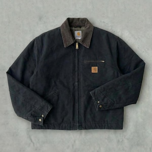 Op de afbeelding: Zwarte werkjas met een bruine corduroy kraag. De jas heeft een ritssluiting, meerdere zakken en een kleine vierkante logopatch op de borst. De mouwen hebben manchetten met knopen.