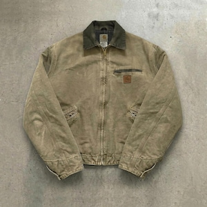 May include: A Carhartt work jacket in a faded olive green colour. The jacket has a dark green collar, a zip closure, and multiple pockets. A small Carhartt logo patch is visible on the chest. The jacket appears to be made of a durable, heavy-duty fabric.