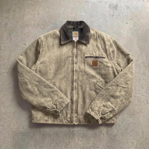 May include: A beige canvas work jacket with a dark grey corduroy collar and a full-length front zip. The jacket features a zipped chest pocket, two lower slanted pockets, and a small brown square logo patch on the left chest area.