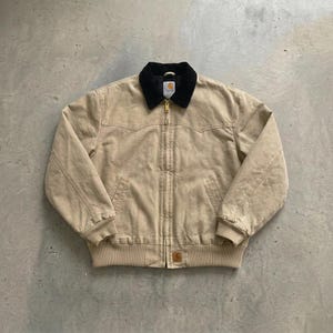 May include: A tan canvas jacket featuring a black corduroy collar, a full-length front zip, and ribbed cuffs and hem. The garment includes a Carhartt brand label on the chest and a small square logo patch on the lower left front pocket area.