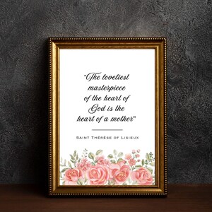 May include: Framed print with a gold-colored ornate frame. The print features the quote "The loveliest masterpiece of the heart of God is the heart of a mother" by Saint Thérèse of Lisieux, with pink floral accents.