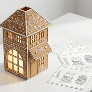 May include: A gingerbread house with intricate white icing details, including snowflakes and swirls, sits on a white surface. The house has multiple windows and a small door. Architectural diagrams are next to the house.