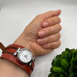 May include: A wristwatch with a silver case and a white face, featuring Roman numerals, is attached to a wide, wrap-around, reddish-brown leather band. The watch is on a wrist, with a hand and green foliage in the background.