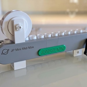 May include: A gray and white mechanical device with a hand crank, featuring a large white wheel and a row of white cylindrical components. The side is printed with the text JP Mini Midi Max and a planet icon, with a green JUNIOR MINI label attached.