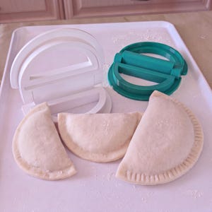 May include: A white plastic dumpling press and a teal plastic dumpling press are positioned behind three uncooked, semi-circular dough pockets. The dough pockets feature crimped edges, and the surface is lightly dusted with flour.