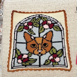 Puede incluir: Cojín cuadrado de ganchillo con un diseño de gato en estilo vidriera. El gato es naranja y marrón, rodeado de hojas verdes y flores rosas y rojas. El borde es crema y marrón.