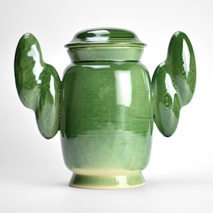 May include: A green ceramic jar with a lid and two wing-like handles. The jar has a glossy finish and a rounded shape, with a lighter green base. The design is modern and artistic, suitable for home decor.
