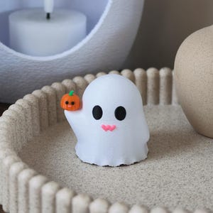 Op de afbeelding: Een klein wit spookfiguurtje met grote zwarte ogen en twee roze hartvormige wangen. Het spookje heeft één arm omhoog en houdt een piepkleine oranje pompoen vast met een groen steeltje en een klein zwart gezichtje op het oppervlak.
