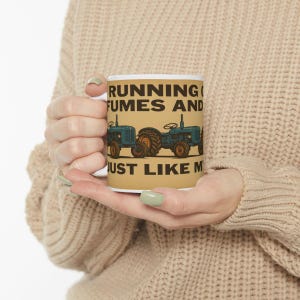 May include: White ceramic mug with the text "RUNNING FUMES AND JUST LIKE M" in black. The mug features two blue tractors with orange wheels on a tan background. The mug is held by a person wearing a beige sweater.