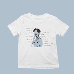 Pode incluir: Uma t-shirt branca de manga curta com um gráfico central de uma pessoa com um casaco com padrão de estrelas. O design inclui uma lista de faixas com títulos como Hit the Wall, Death Wish, The Knife, Daughter from Hell e Cold Goodbyes.