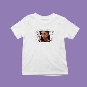 Pode incluir: T-shirt branco com um retrato de Gracie Abrams em uma moldura decorativa. O texto "GRACIE ABRAMS" está acima da imagem e "A DAUGHTER FROM HELL" abaixo. A camiseta é exibida em um fundo roxo.