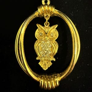 May include: A gold-toned owl pendant with a rhinestone-studded body hangs within a gold oval frame. The owl's eyes are also adorned with rhinestones. The frame has a textured design, and the pendant is suspended from a small loop.