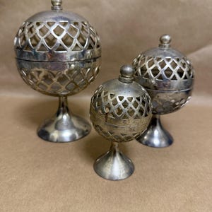 May include: Three silver-colored, spherical incense burners with intricate lattice designs. Each burner has a pedestal base and a removable lid with a small knob. The burners vary in size and are displayed on a neutral-toned surface.