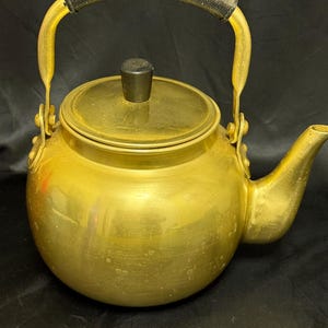 May include: A gold-colored metal teapot with a black handle and lid knob. The teapot has a rounded body, a spout, and a hinged handle. The handle has a black, textured grip. The teapot is likely used for boiling water.