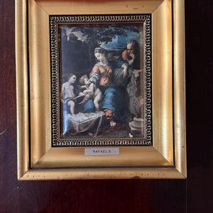 May include: A rectangular gold-toned frame containing a reproduction of a classical painting depicting a family group in a landscape. The frame features an inner decorative border and a small silver plaque at the bottom with the text RAFAEL S.
