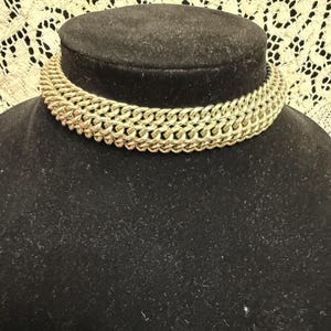 May include: A gold-toned chain necklace with a thick, woven design. The necklace is displayed on a black velvet bust, set against a cream-colored, lace-like background.