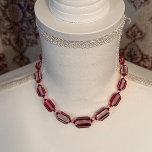 May include: A necklace featuring a series of rectangular, faceted glass beads in alternating clear and deep red stripes. The beads are strung together on a thin red cord, creating a uniform pattern that encircles the neck in a single strand.