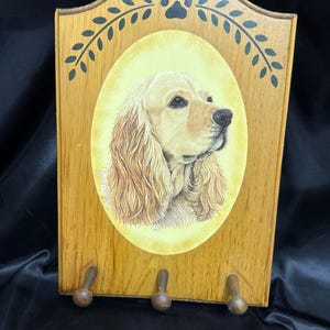 May include: Wooden key holder featuring a golden Cocker Spaniel portrait in an oval frame. The key holder has three wooden pegs and is decorated with paw prints and leaf accents. The wood has a light brown finish.
