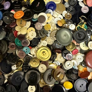 May include: A collection of various buttons in different sizes, shapes, and colors. The buttons include black, white, gold, and other colors. Some buttons have two holes, while others have four. The buttons are made of plastic and metal.