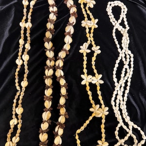 May include: Four shell necklaces of varying designs. One necklace features small, round shells, while another has larger, oval shells with brown accents. A third necklace has flower-shaped shells, and the last is made of small, white shells.