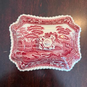 May include: A rectangular ceramic butter dish lid with a scalloped edge, featuring a red and white transferware pattern of a landscape with trees, a stone bridge, and a castle. A square knob with a floral design sits in the center of the lid.