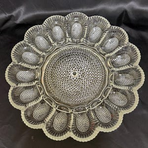 May include: Clear glass serving dish with a scalloped edge and a flower-like design. The dish has a central circular area with a textured pattern and individual egg-shaped compartments around the perimeter. The glass has a textured, detailed surface.