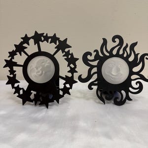May include: Two decorative metal objects with black frames and frosted glass centers. One frame features a circular arrangement of stars surrounding a moon face, while the other features swirling sun rays surrounding a sun face with facial features.