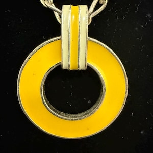 May include: A vintage necklace featuring a bright yellow enamel pendant in a circular shape. The pendant is accented with a white and yellow bar, and hangs from a silver-toned chain. The jewelry is set against a black background.
