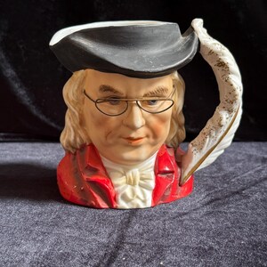 May include: A ceramic character jug depicting a person wearing a black tricorn hat, wire-rimmed glasses, and a red coat with a white cravat. The handle of the jug is shaped like a white quill pen with gold-colored accents on the textured surface.