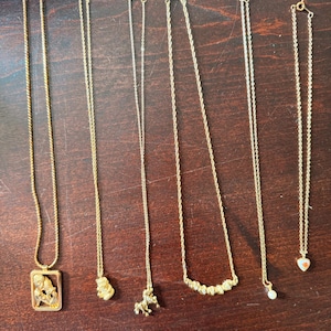 May include: Six gold-toned necklaces arranged vertically. The first features a rectangular pendant with a figure, the second a small charm, the third a horse-shaped charm, the fourth a curved bar with textured accents, the fifth a single pearl, and the sixth a small heart charm.