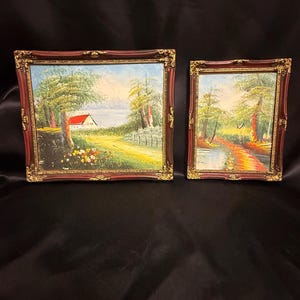 May include: Two framed landscape oil paintings. The larger painting depicts a house with a red roof, trees, and a body of water. The smaller painting shows a path through trees. Both paintings have ornate, dark red frames with gold accents.