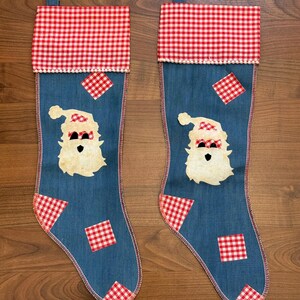 May include: Two blue denim Christmas stockings featuring a red and white gingham cuff and matching patches. Each stocking displays a central applique of a Santa Claus face with a gingham hat and eyes, finished with a small white pom-pom trim.