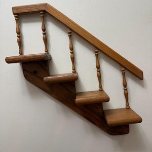 May include: A wall-mounted wooden shelf shaped like a miniature staircase with four steps, a diagonal handrail, and vertical turned spindles. The wood has a medium brown finish with visible grain patterns, featuring a sturdy side support structure.