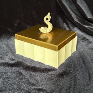 May include: A rectangular box with a cream-colored, vertically fluted base and a smooth, metallic bronze-toned lid. A sculpted, light-colored dolphin figure serves as a handle on top of the lid, positioned centrally to allow for easy lifting.