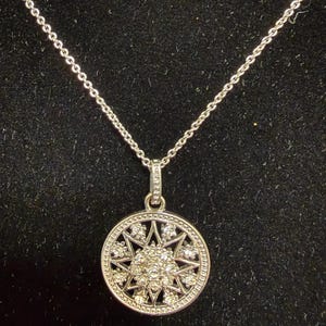 May include: Silver-toned necklace with a circular pendant featuring a starburst design. The pendant is adorned with small, sparkling stones and hangs from a delicate chain. The necklace is displayed against a dark background.