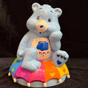 May include: A light blue ceramic figurine of a bear sitting on a colorful, multi-paneled base. The bear has a white belly patch featuring a dark blue rain cloud with three red hearts and raindrops. The base is decorated with small hearts.