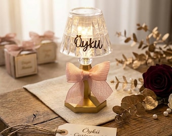 Op de afbeelding: Een kleine, decoratieve mini-lamp met een heldere, facetgeslepen lampenkap. De lamp heeft een gouden voet en een roze strik. De tekst "Öykü" staat op de lampenkap. Een label leest "Öykü - Customizable Mini Lamp".