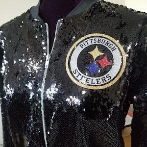 Puede incluir: Chaqueta bomber de lentejuelas negras con cremallera plateada y parche con el logotipo de los Steelers de Pittsburgh. El parche presenta el nombre y el logotipo del equipo en dorado, plateado, rojo, azul y amarillo.