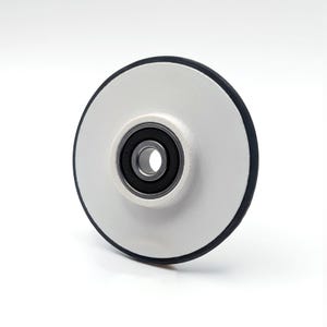 May include: A circular wheel featuring a smooth white disc surface with a dark gray outer rim. The center of the wheel contains a metallic ball bearing assembly with a dark inner ring, designed for rotational movement and mechanical applications.
