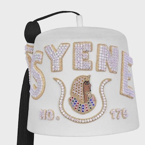 May include: White fez hat with the word "SYENE" in sparkling letters. The hat features an embroidered design of an Egyptian pharaoh and the numbers "176". A black tassel hangs from the side.