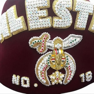 Puede incluir: Un fez burdeos con las palabras "MEST" y "NO. 18" bordadas en blanco, dorado y cuentas iridiscentes. El sombrero presenta un emblema adornado con un diseño de faraón dorado y una estrella.