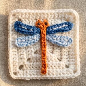 May include: A square crochet motif in off-white yarn featuring a central dragonfly appliqué. The dragonfly has an orange body, dark blue upper wings, and light blue lower wings with small black bead eyes attached to the head section.